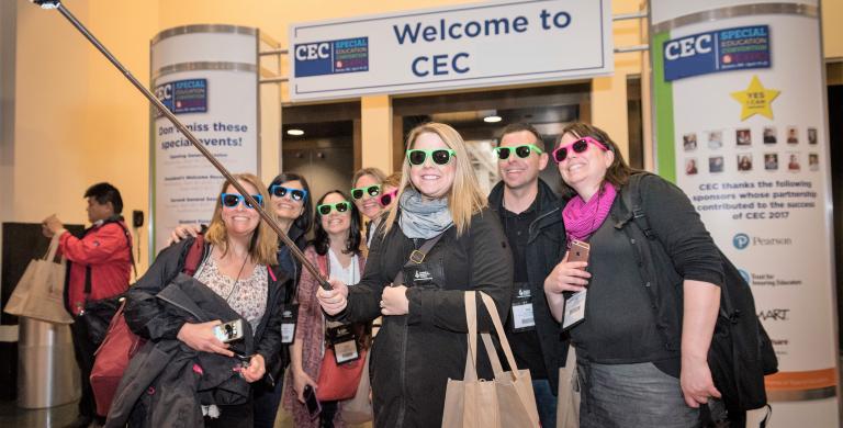 Large group of female members wearing sunglasses and using a stick to take a selfie outside of the Convention Expo Hall